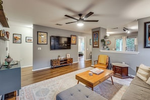 A spacious living room with hardwood flooring, a ceiling fan, a flat-screen TV, a wooden coffee table, a grey sofa, and a patterned rug.
