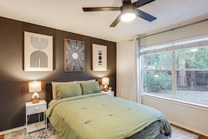 A bedroom with a bed, bedside tables, wall art, a ceiling fan, and a window with a view of greenery. - Easy Airport Access! Austin Home w/ Patio (Austin)