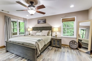 Quarto com cama, ventoinha no teto, mesa de cabeceira, espelho e janela com cortinas.