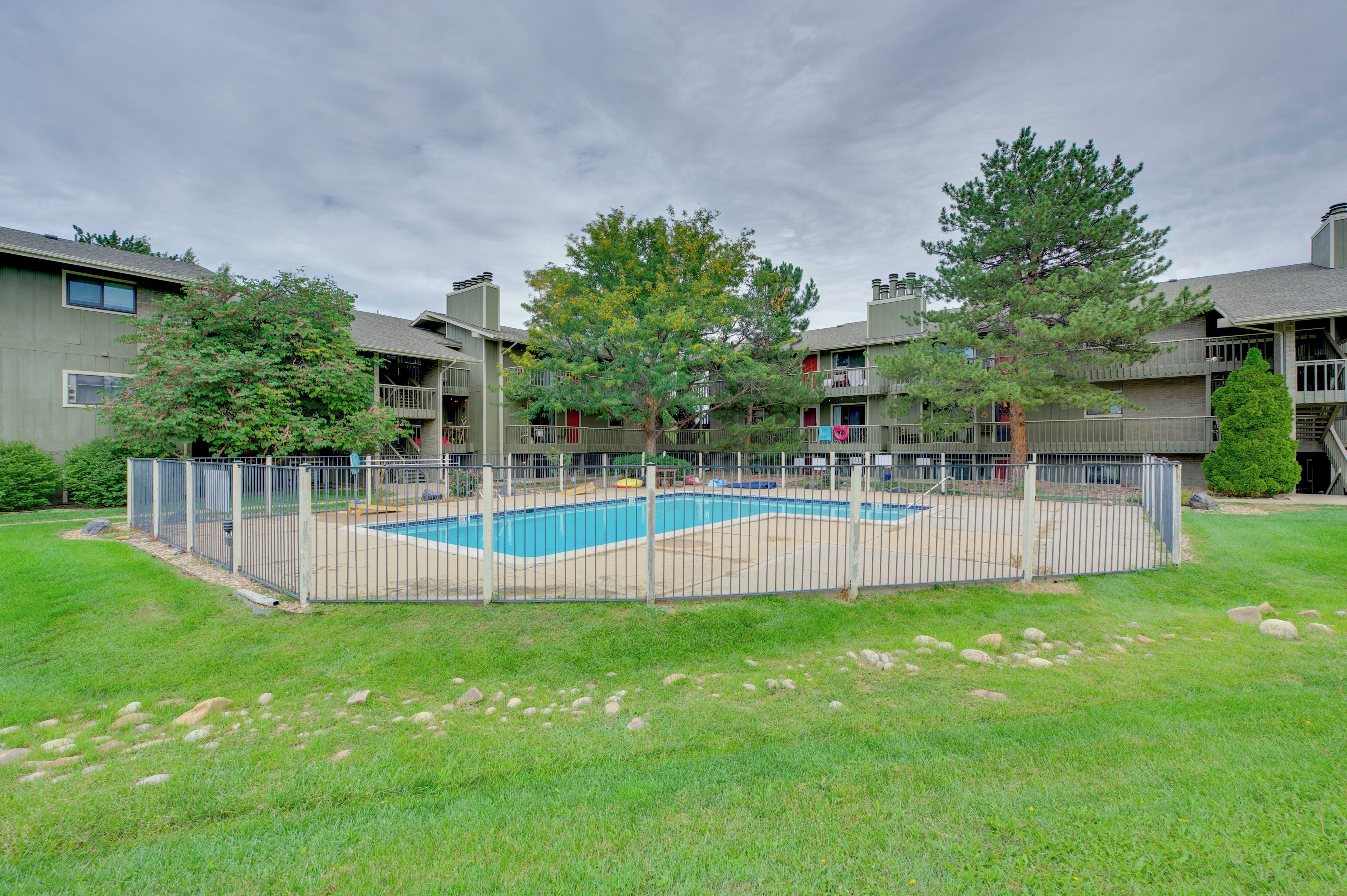 A residential area with a swimming pool surrounded by a fence, trees, and a grassy lawn.