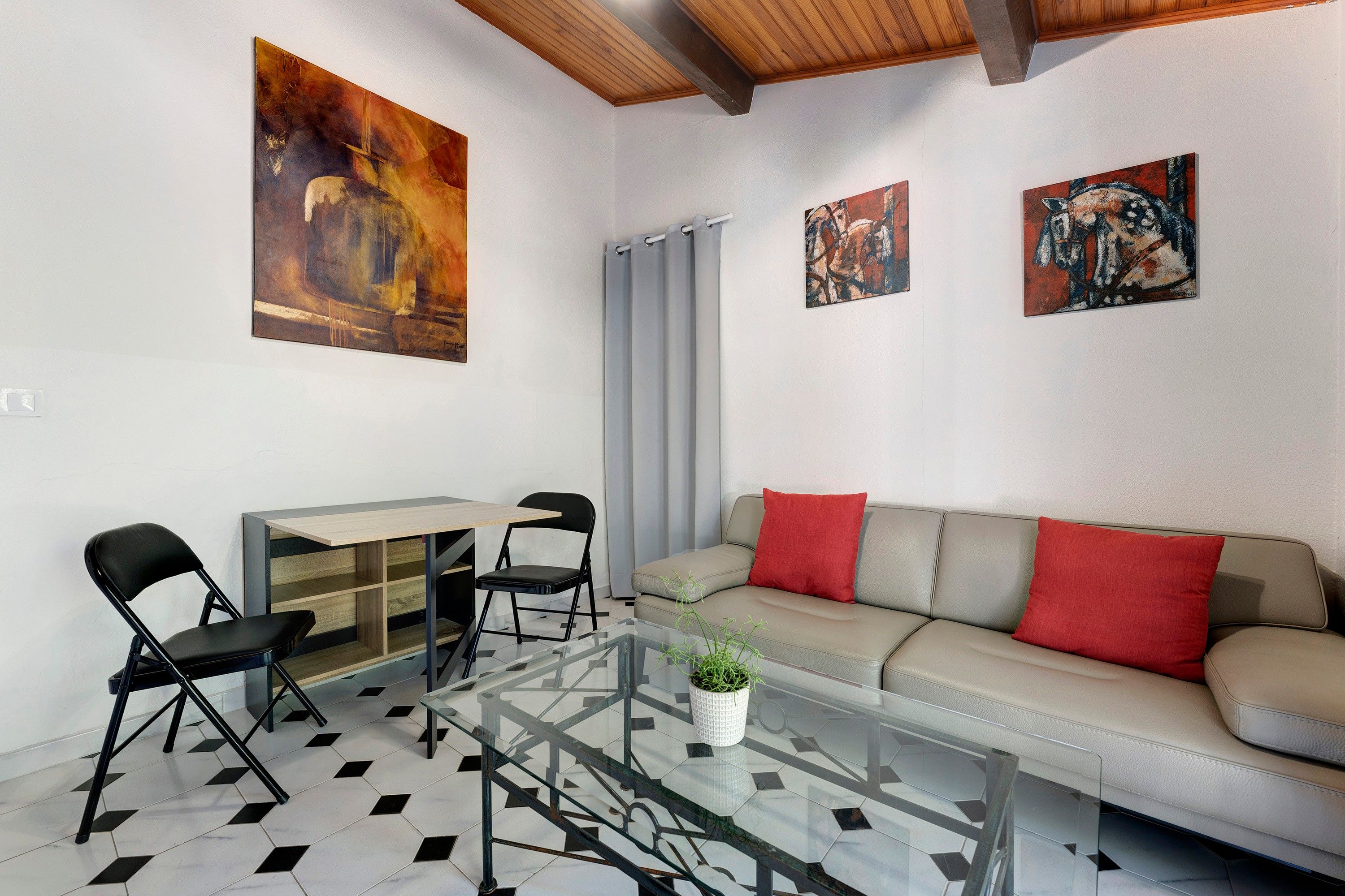 Un salon moderne avec une table basse en verre, un canapé avec des coussins rouges, un coin repas avec une table et des chaises, et des œuvres d’art accrochées aux murs.