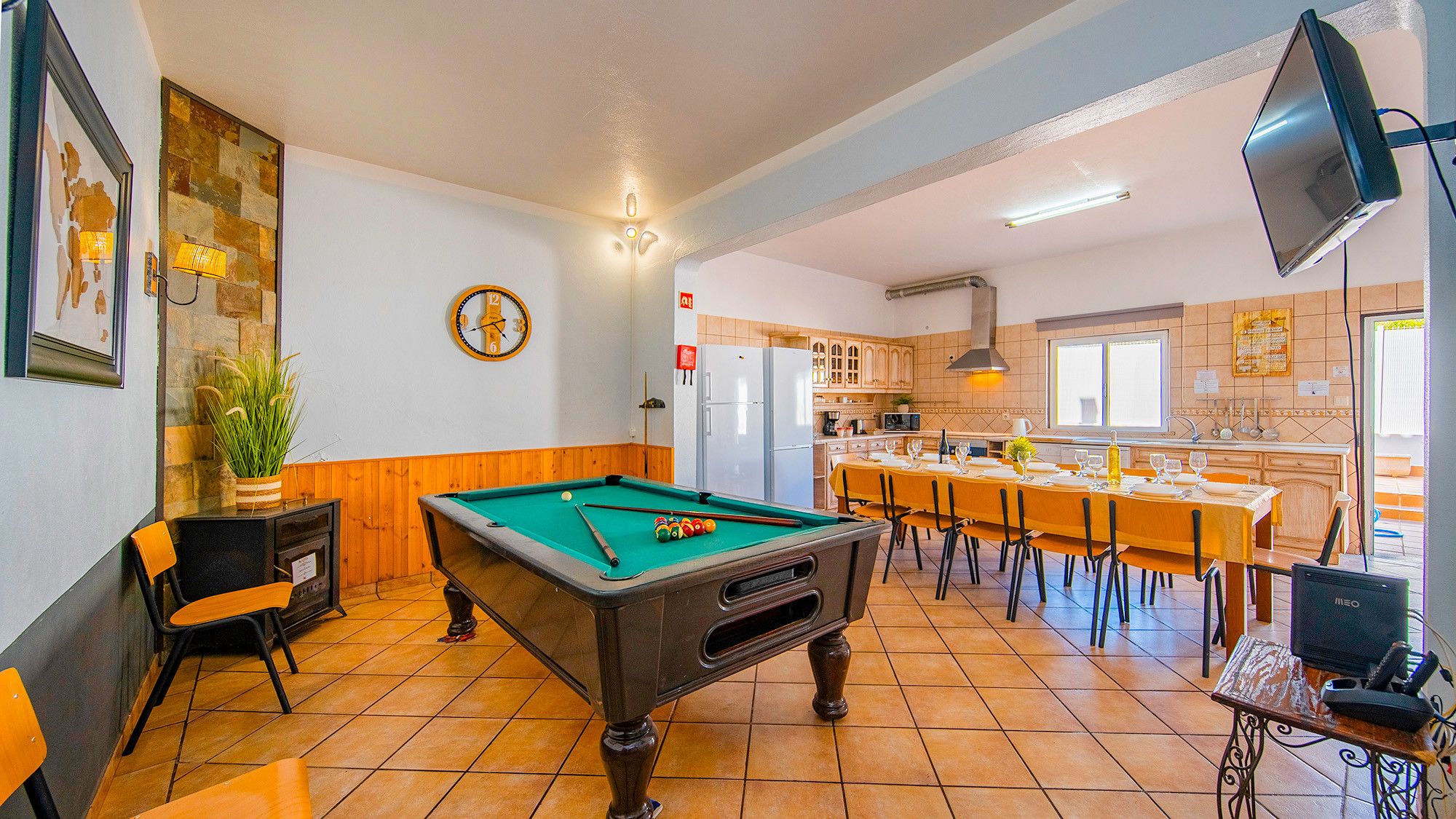 A pool table in a room with a kitchen area, wooden flooring, and a clock on the wall.