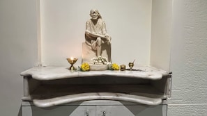 Una estatua de mármol de una figura sentada sobre un pedestal, con una vela encendida, flores y un objeto de bronce sobre una base de mármol blanco.