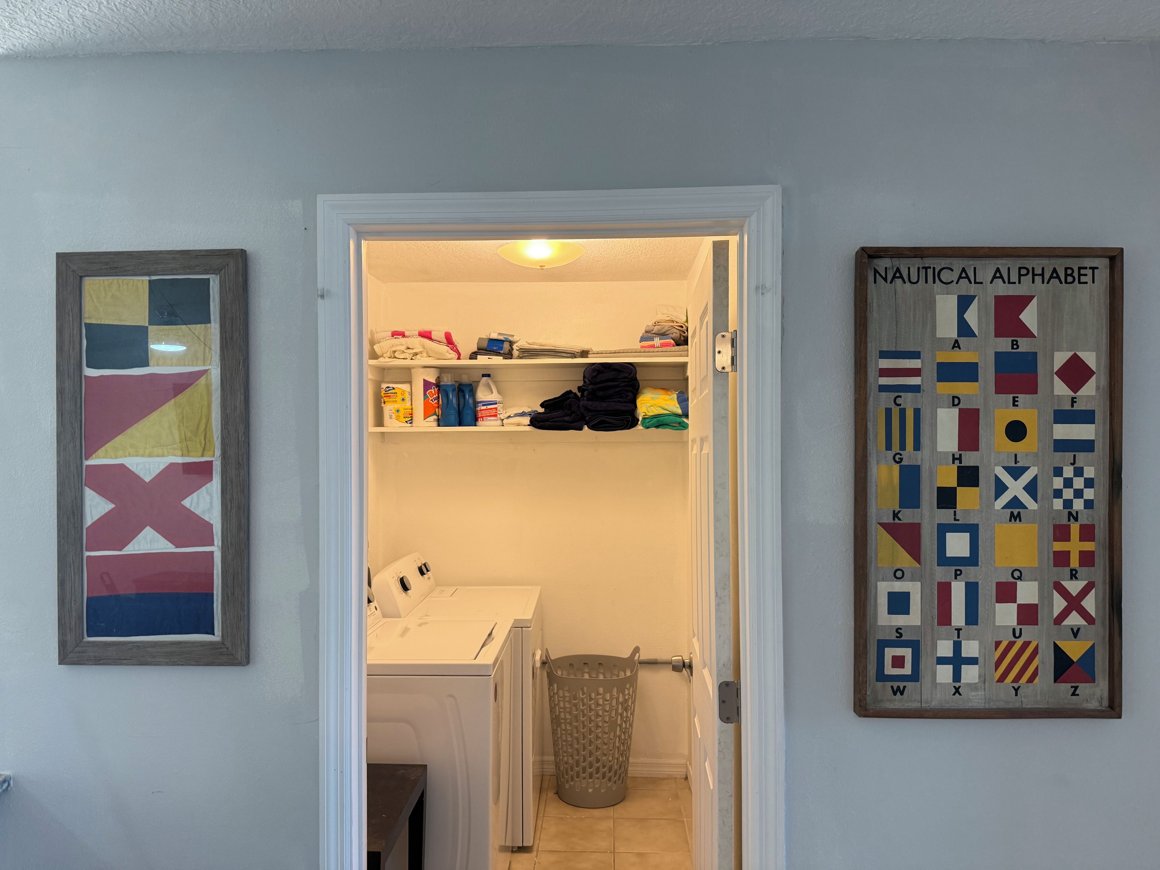 Ett tvättrum med tvättmaskin, torktumlare och en papperskorg. På väggen hänger en tavla med ett marint alfabet och en färgstark, abstrakt målning.