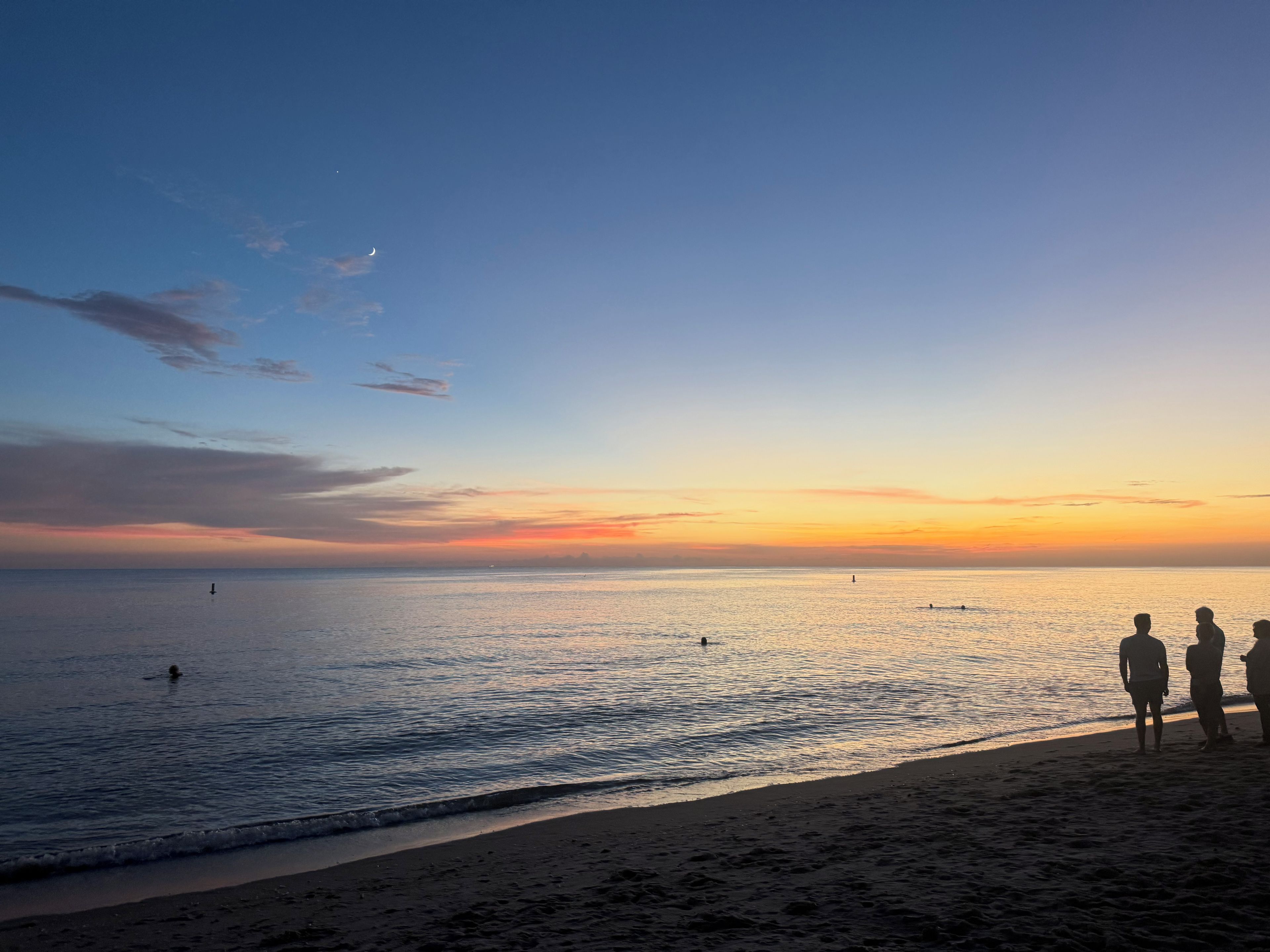 A beach with a sunset, calm sea, and silhouettes of people.