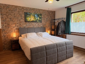 A bedroom with a stone wall, a bed with a grey upholstered headboard, a hanging light fixture, a window with curtains, and a coat rack with clothes. - Spessart Dream: 17 guests, 5 bedrooms, foosball, and a view of the valley in the nature park (Sinntal)