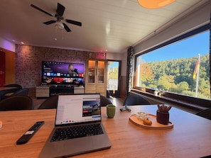 A modern room with a laptop, a TV displaying a streaming service, and a large window with a view of trees. - Spessart Dream: 17 guests, 5 bedrooms, foosball, and a view of the valley in the nature park (Sinntal)