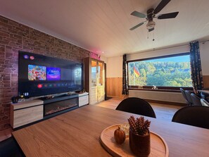 A modern living room with a flat-screen TV, wooden dining table, and a view of a forest through a large window. - Spessart Dream: 17 guests, 5 bedrooms, foosball, and a view of the valley in the nature park (Sinntal)