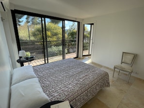 A bedroom with a large bed, a chair, a nightstand, and a view of a balcony with a garden. - Villa Grand Bleu (La Seyne-sur-Mer)