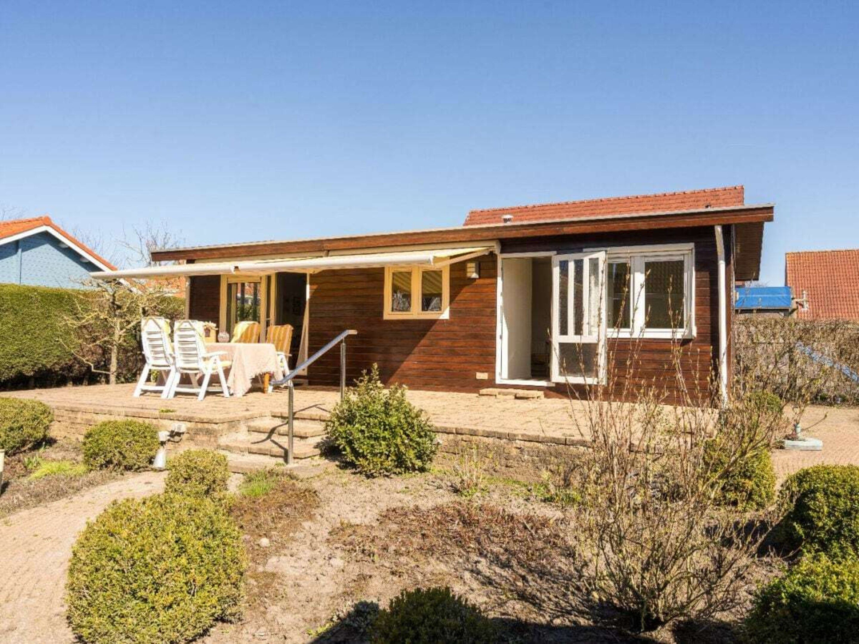 A single-story wooden house with a covered patio, a small garden, and a clear sky.