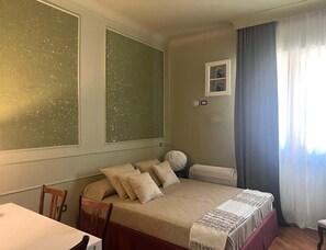 Ein Schlafzimmer mit Bett, Stuhl, Tisch, Heizkörper und einem fenster mit Vorhängen.