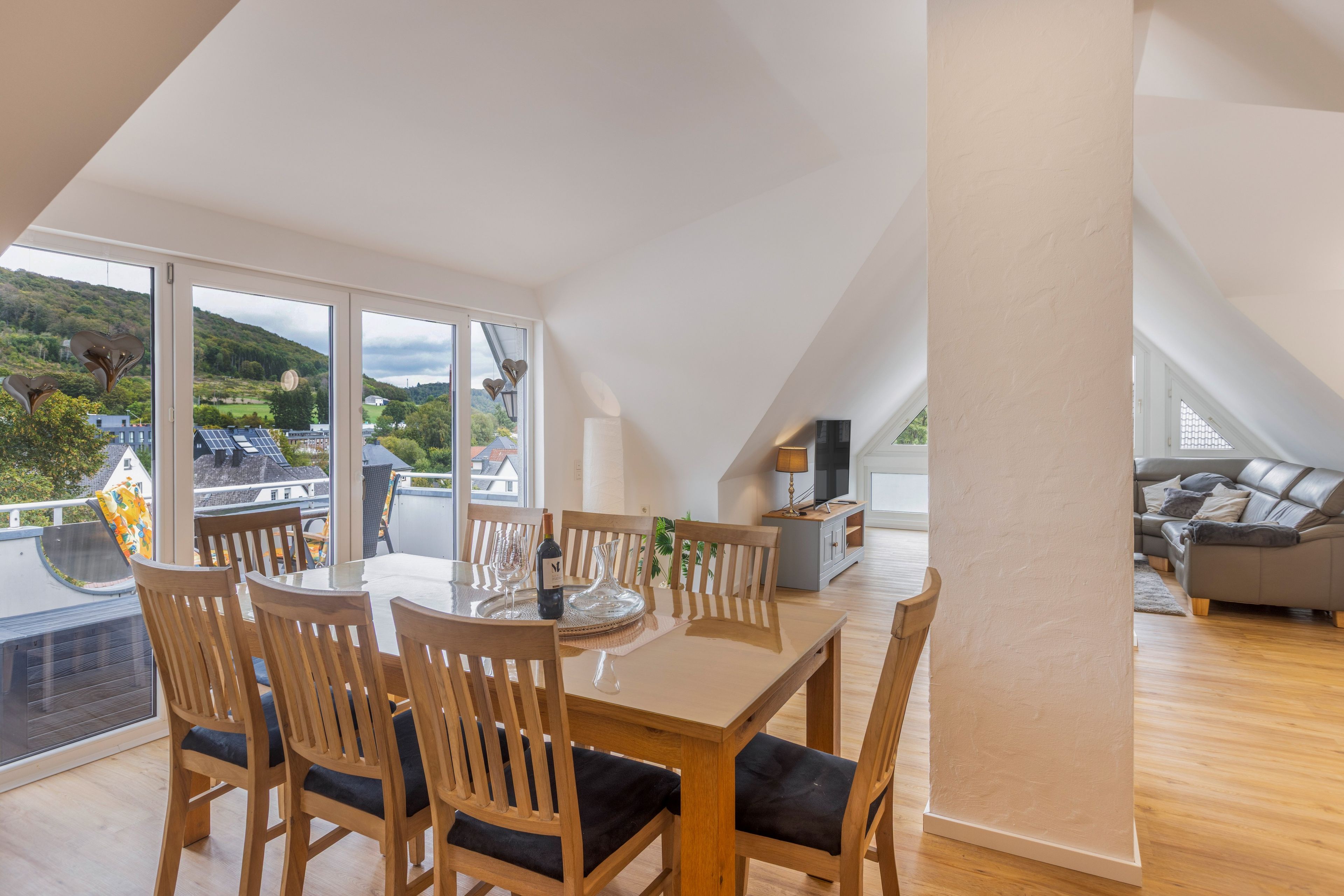Un coin repas avec une table dressée pour quatre personnes, des chaises en bois et une vue sur un paysage verdoyant grâce à de grandes fenêtres.
