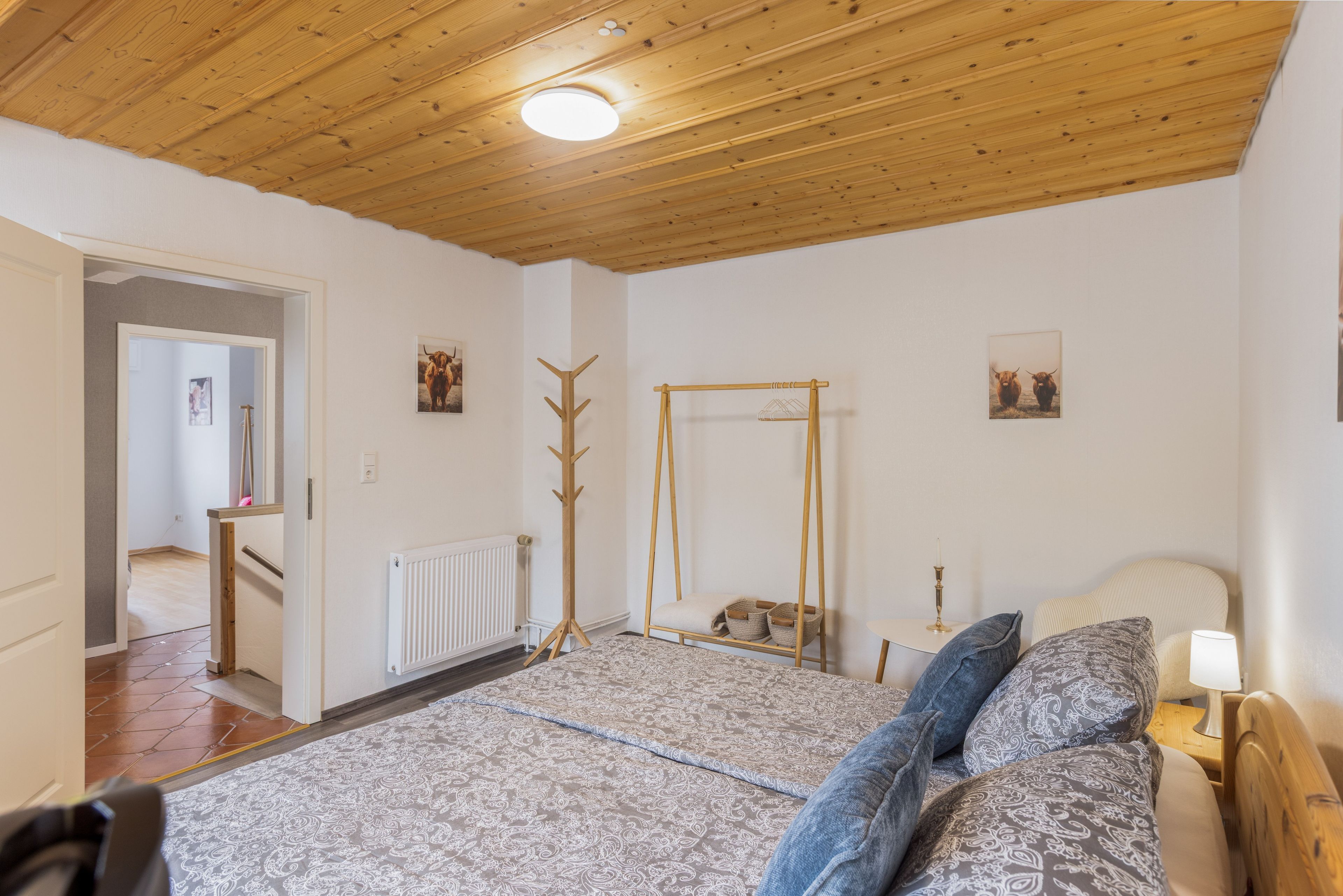 Une chambre avec un plafond en bois, un lit avec une literie à motifs, une table de chevet avec une lampe et un porte-manteau.