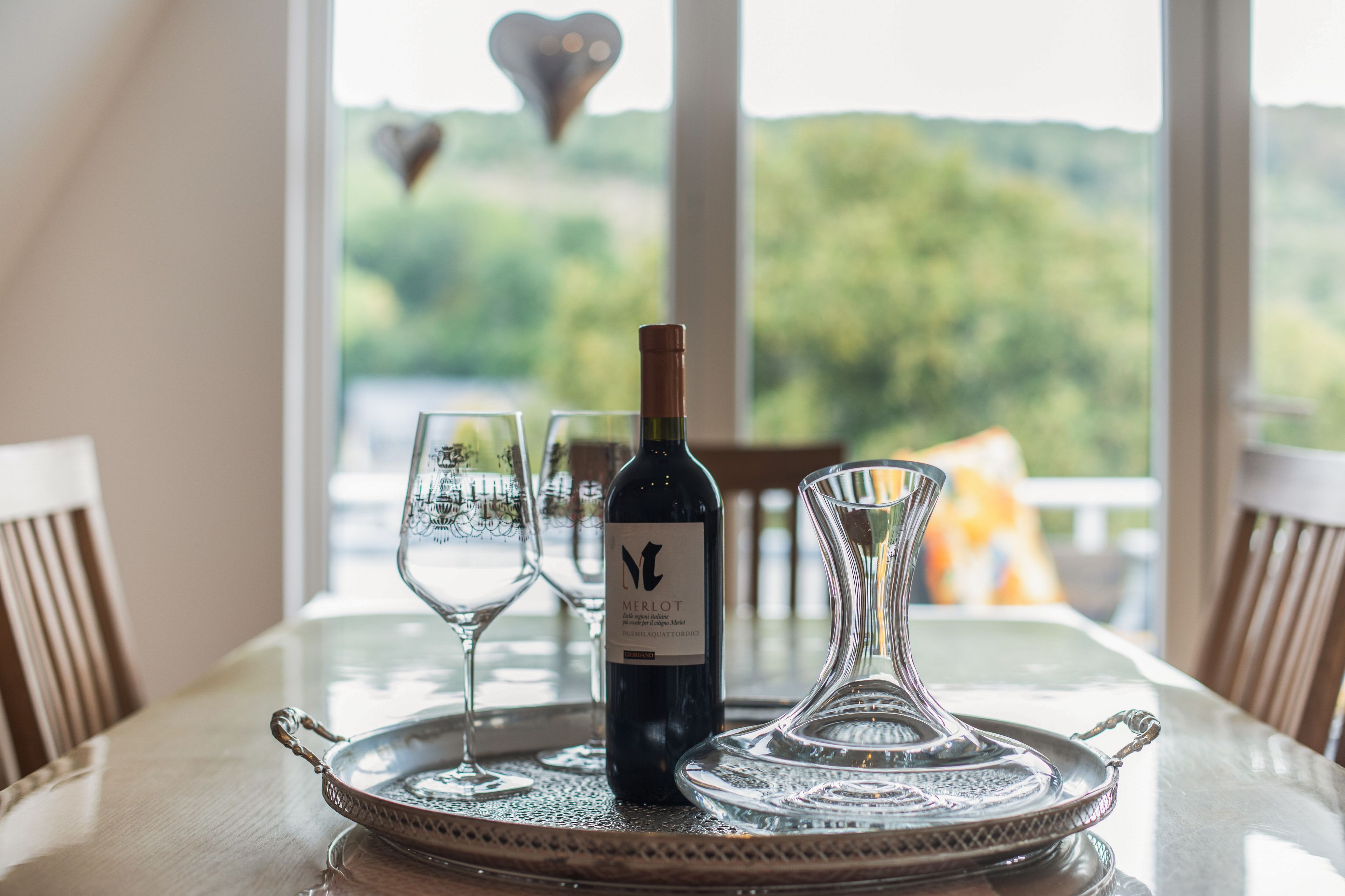 Eine Flasche Merlot Wein auf einem Tablett zusammen mit zwei Gläsern und einer Karaffe auf einem Tisch, mit Blick auf Bäume durch ein Fenster.