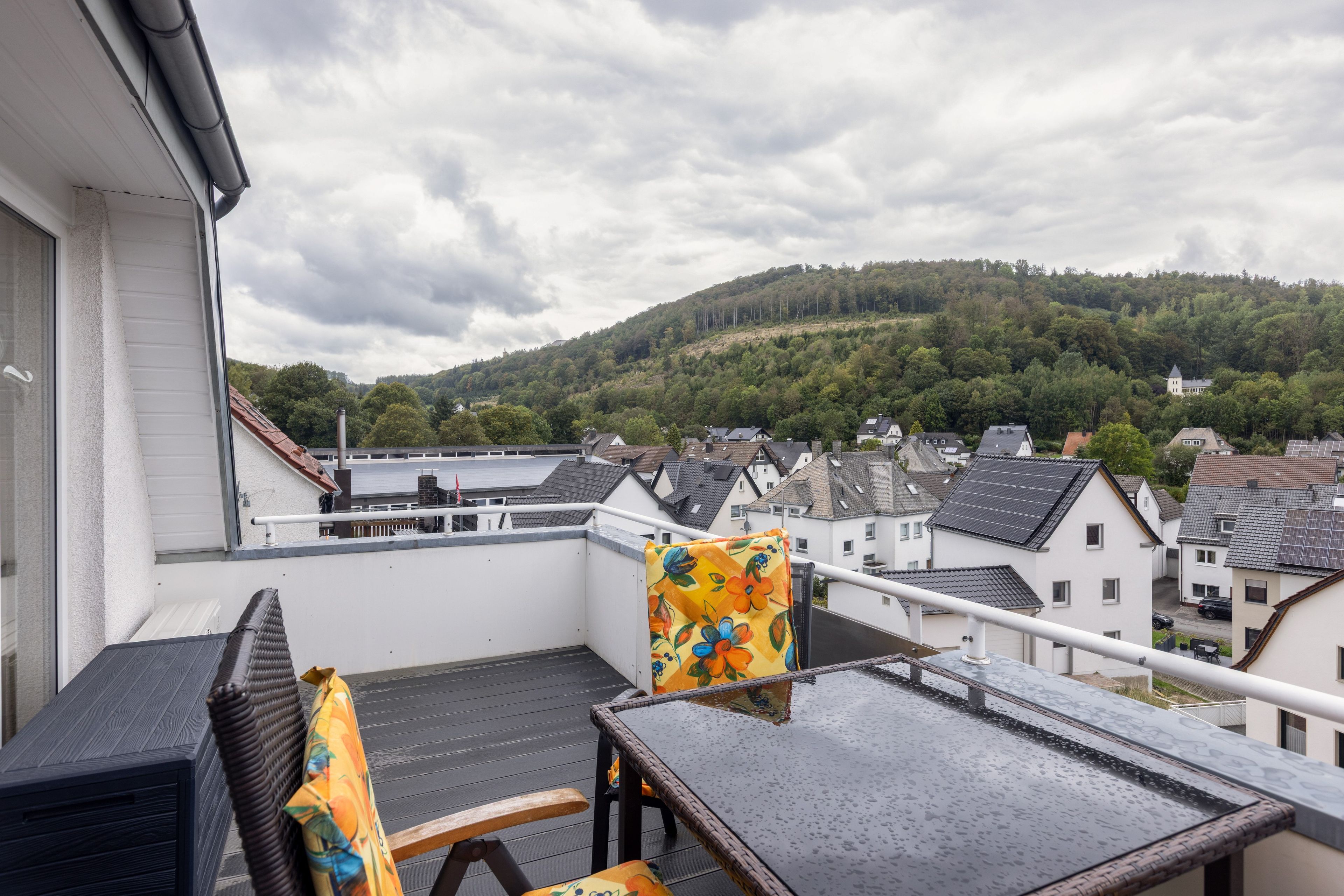 Eine Dachterrasse mit Blick auf einen bewaldeten Hügel, ein weißes Gebäude und ein schwarzer Stuhl.