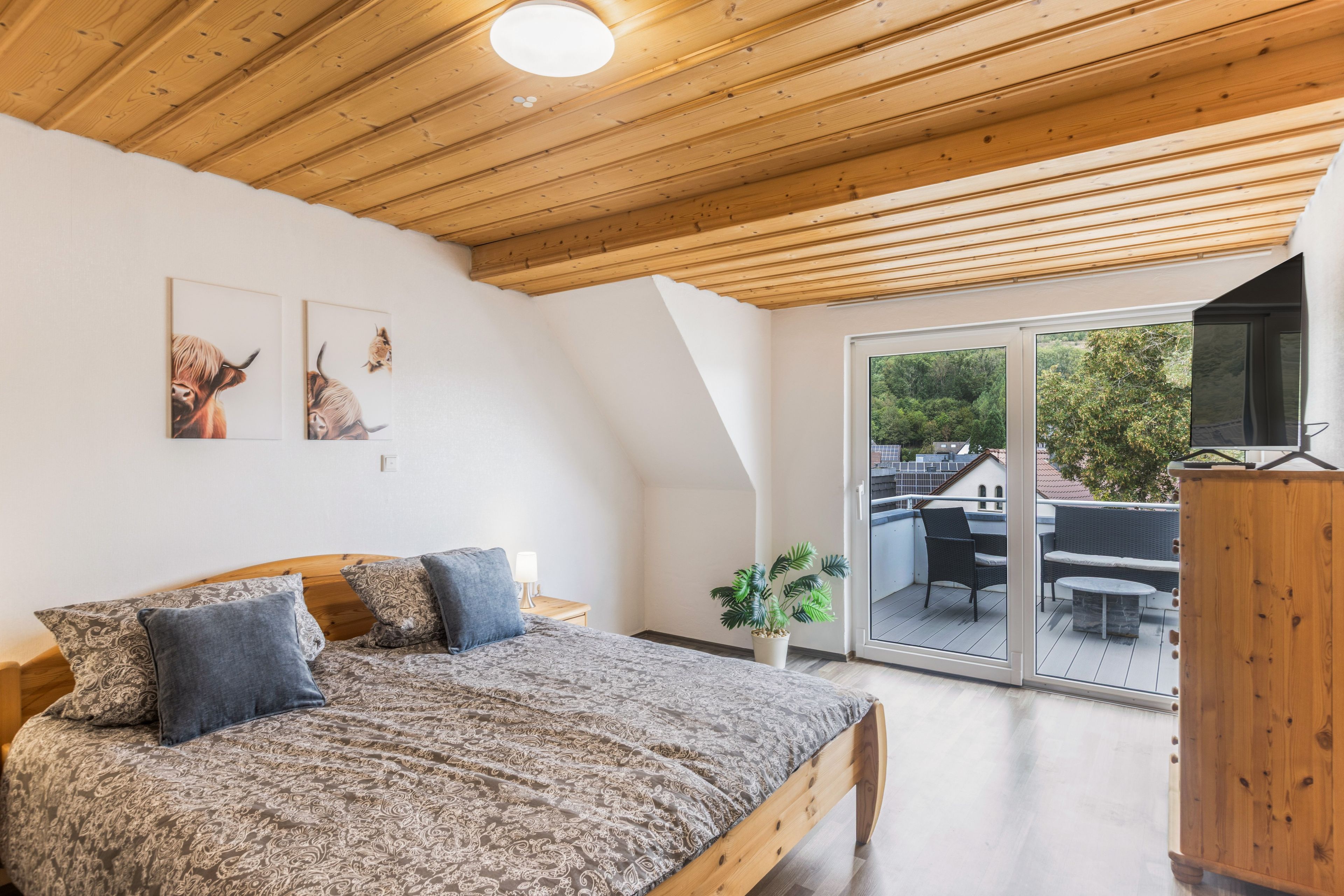 Ein Schlafzimmer mit Holzbalkendecke, einem Bett mit gemusterten Betttextilien, zwei an der Wand befestigten Tierbildern und Blick auf einen Balkon mit Gartenmöbeln.