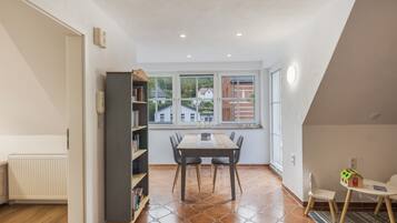 Een zithoek met een tafel en stoelen, een boekenkast en uitzicht op gebouwen door een raam.