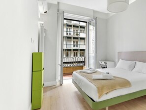 Ein Schlafzimmer mit einem Bett, einem grünen Schrank, einem Schreibtisch und Blick auf ein Gebäude durch eine offene Tür.