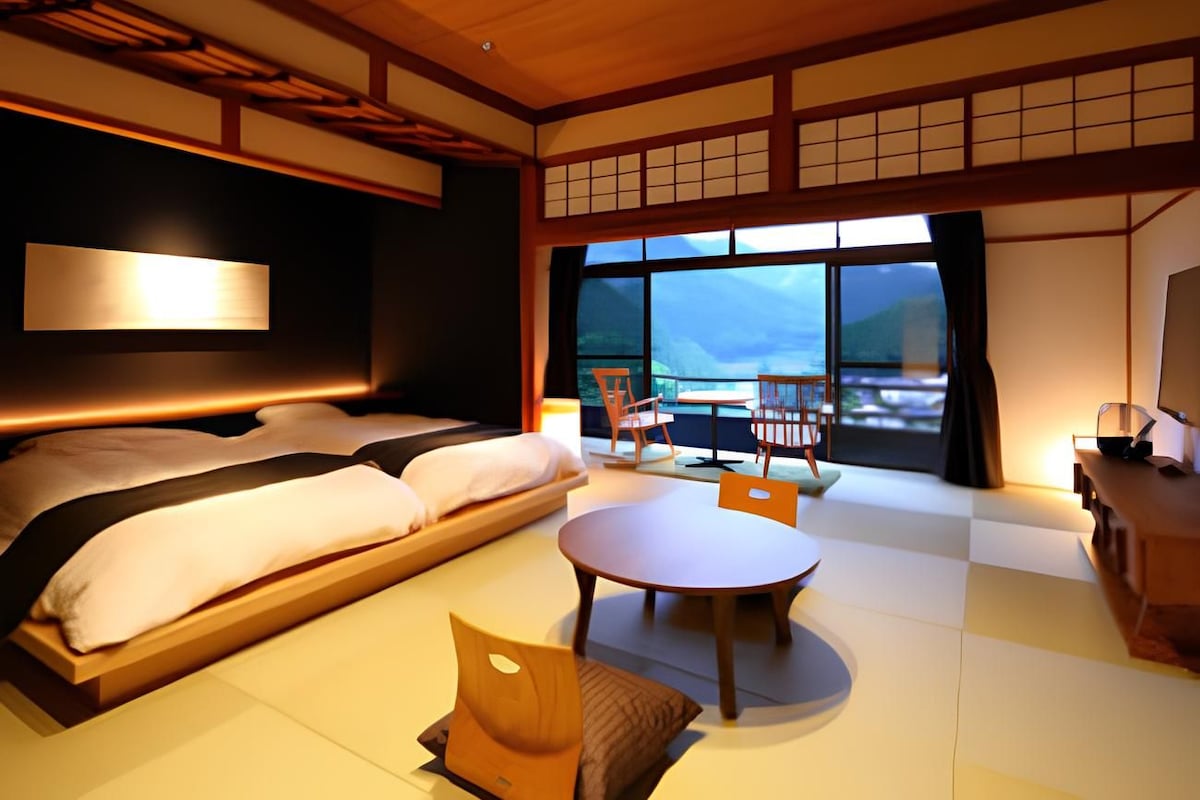 Habitación de hotel con cama, mesa de comedor, sillas, televisor y un ventanal con vistas.