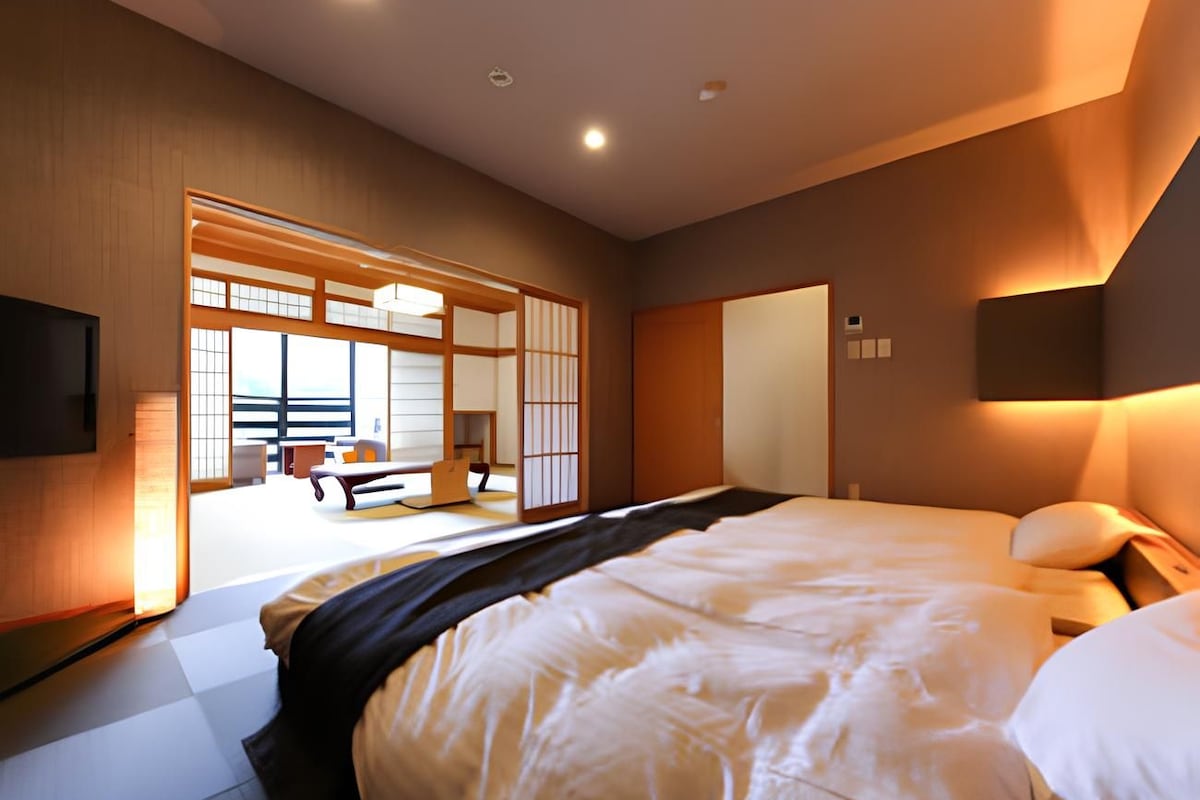 Habitación de hotel con una cama grande, televisión, puerta corredera que da a una habitación tradicional japonesa y mesita de noche con lámpara.