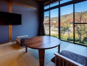 A room with a round wooden table, a bench, tatami flooring, a TV mounted on the wall, and a large window with a view of hills. - Furuyu Onsen Ryokan Suginoya (Saga)