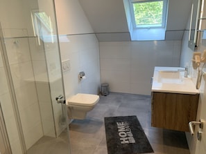 A modern bathroom with a glass shower enclosure, a toilet, a bidet, and a sink with a wooden vanity. - Villa Trisatus on the Lake (Goehren-Lebbin)