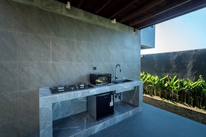 Une cuisine extérieure équipée d’une cuisinière à gaz, d’un four à micro-ondes et d’un évier, installée contre un mur en béton et offrant une vue sur un jardin.
