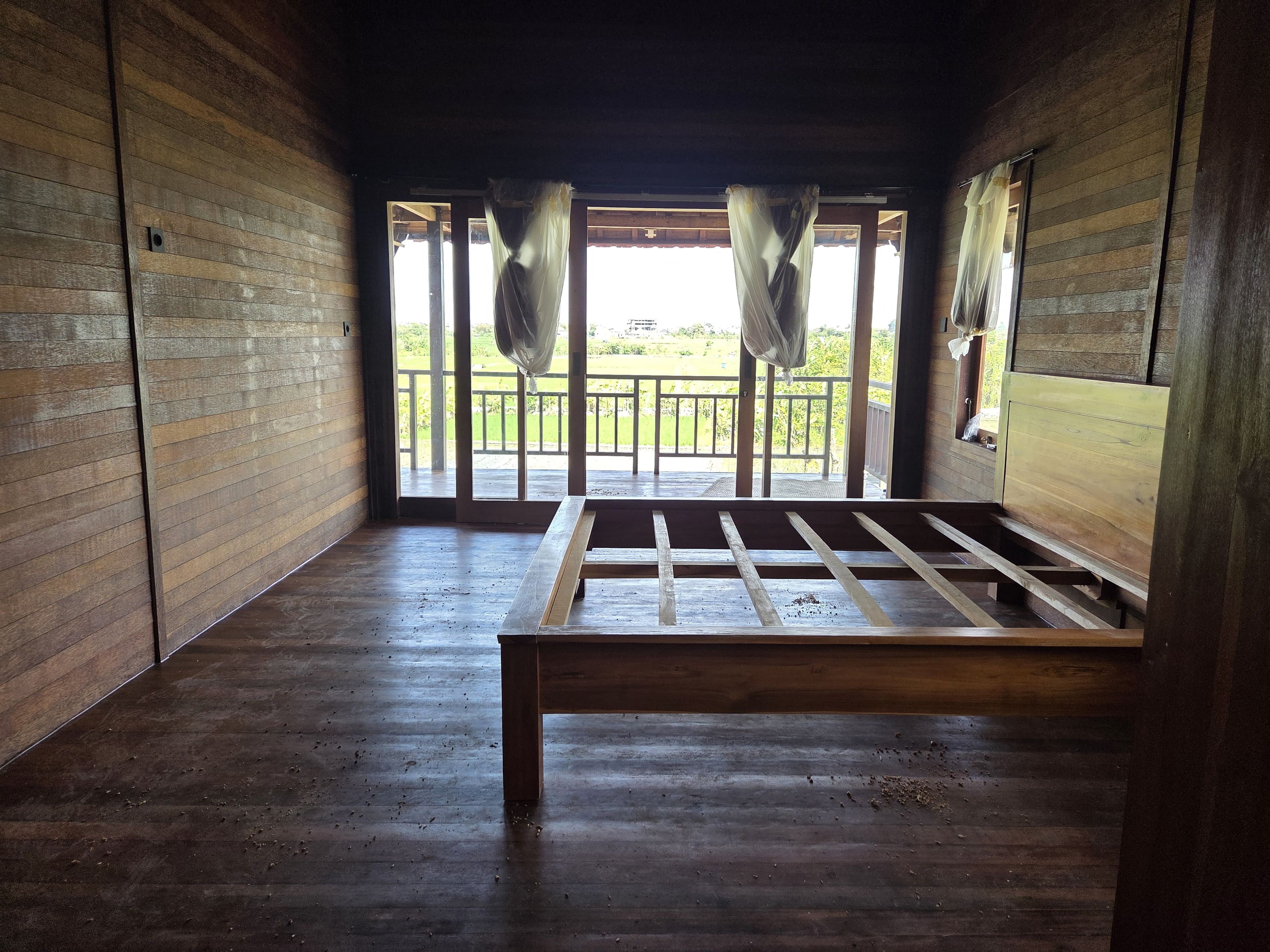 A room with wooden walls and a bed with a metal frame. There is a balcony with a railing and a view of greenery outside.