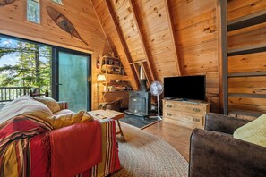 A cozy loft bedroom with a wooden sloped ceiling, a large window, a fireplace, a television, and a cozy seating area with a striped blanket. - Treetop Cabin | Waterfront on Mooselookmeguntic | Pet Friendly (Oquossoc)