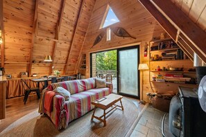 A cozy cabin interior with a wooden ceiling, a sofa, a dining table, and a fireplace. - Treetop Cabin | Waterfront on Mooselookmeguntic | Pet Friendly (Oquossoc)