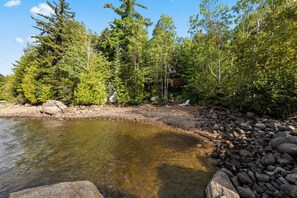 A natural landscape with a clear water body, surrounded by trees and rocks. - Treetop Cabin | Waterfront on Mooselookmeguntic | Pet Friendly (Oquossoc)