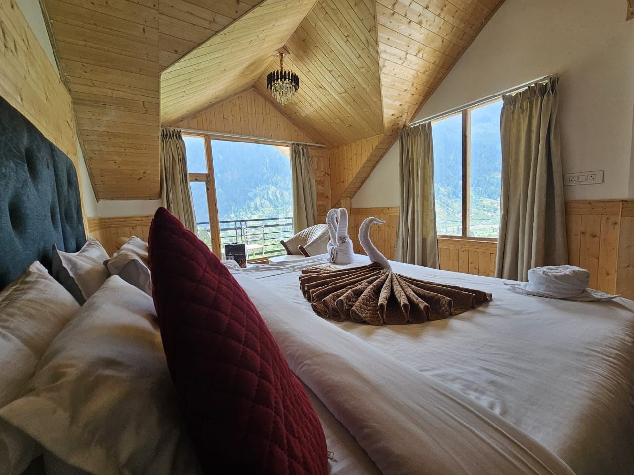A hotel room with a high ceiling, wooden beams, a bed with a red throw pillow, a swan-shaped towel arrangement, and a view of mountains through the windows.