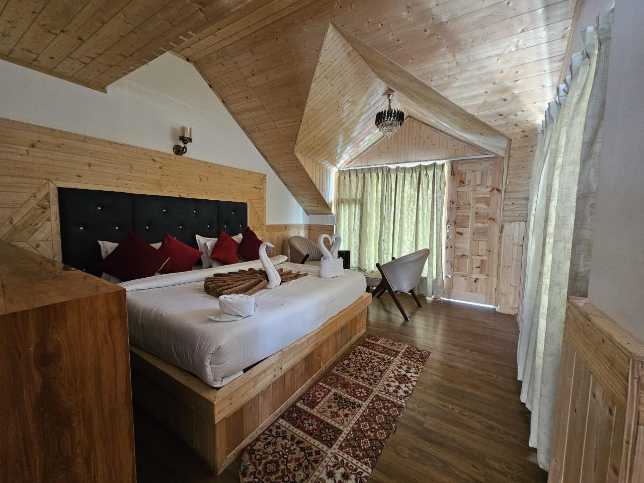 Une chambre avec un grand lit, une tête de lit en bois, un lustre, une chaise et une vue sur l’extérieur.