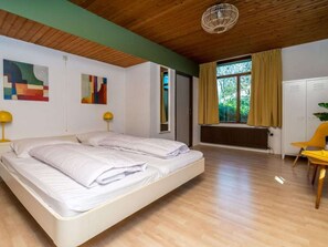 A bedroom with a wooden ceiling, a bed, two bedside lamps, a radiator, a window with curtains, and a chair. - Holiday Home With 4 Bedrooms (Burgh-Haamstede)