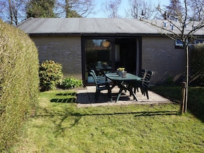 Ein kleines Backsteinhaus mit einer Terrasse, auf der ein Tisch und Stühle stehen, umgeben von einem gepflegten Rasen und einigen Büschen.