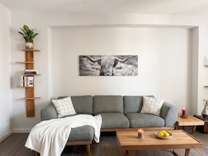 Ein Wohnzimmer mit einem grauen Sofa, einem Couchtisch aus Holz mit Kerze und Zitronen sowie einem an der Wand befestigten Regal mit Büchern und einer Pflanze.