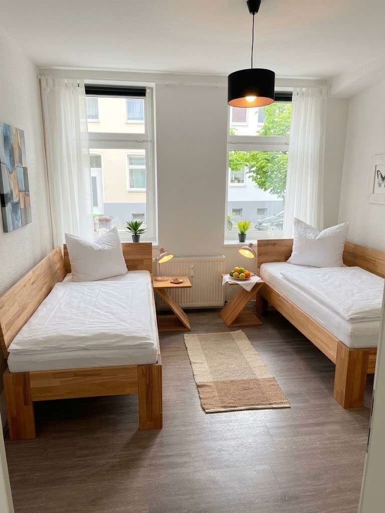Una habitación pequeña y bien iluminada con dos camas de madera, una mesita con una lámpara y vistas al exterior a través de una ventana.