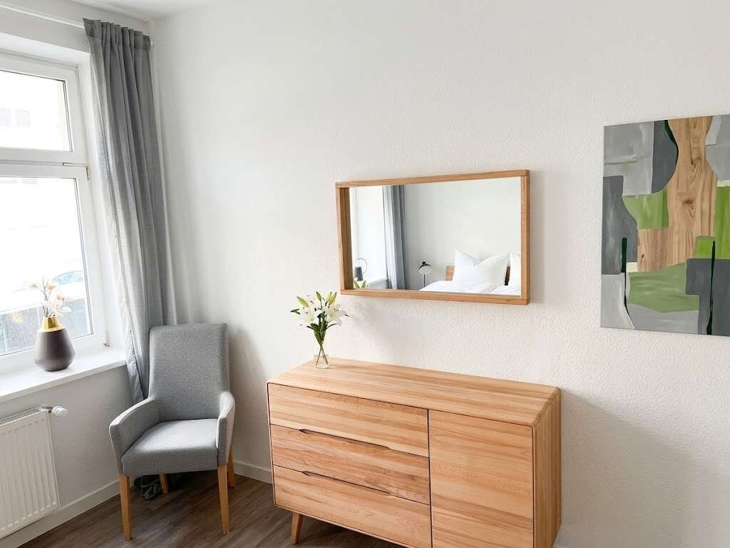 Una habitación con un aparador de madera, un sillón gris, una ventana con cortinas y un espejo en la pared.