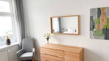 Una habitación con un aparador de madera, un sillón gris, una ventana con cortinas y un espejo en la pared.