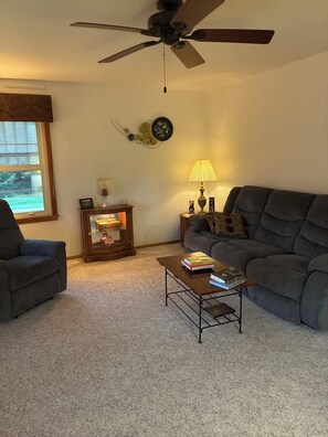A living room with a ceiling fan, a sofa, a coffee table with books, a lamp, and a decorative wall piece. - 2-bedroom farmhouse in soothing  rural Spring Green with AC (Spring Green)