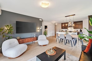 A modern living room with a flat-screen TV, comfortable seating, a dining area with a kitchen in the background. - L'Écrin: Contemporary elegance and cozy comfort in the heart of the city. (Québec)