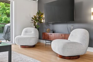 A modern living room with two armchairs, a wooden cabinet, a flat-screen TV, and a large window with a view of greenery. - L'Écrin: Contemporary elegance and cozy comfort in the heart of the city. (Québec)