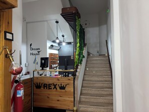 A hotel reception area with a wooden counter, a fire extinguisher, a clock, and a staircase.
