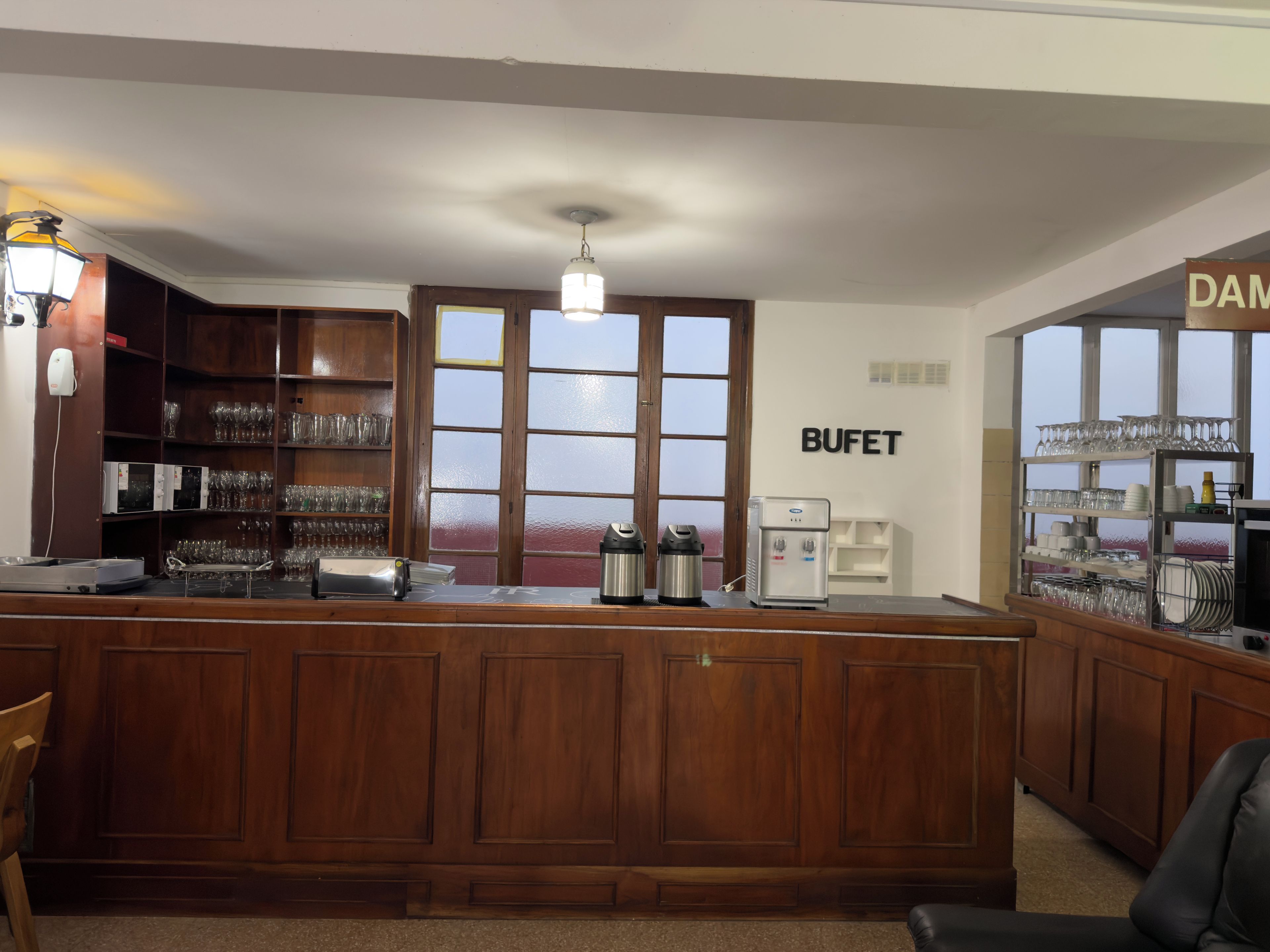 A bar area with a wooden counter, a water dispenser, and shelves with glasses and cups.
