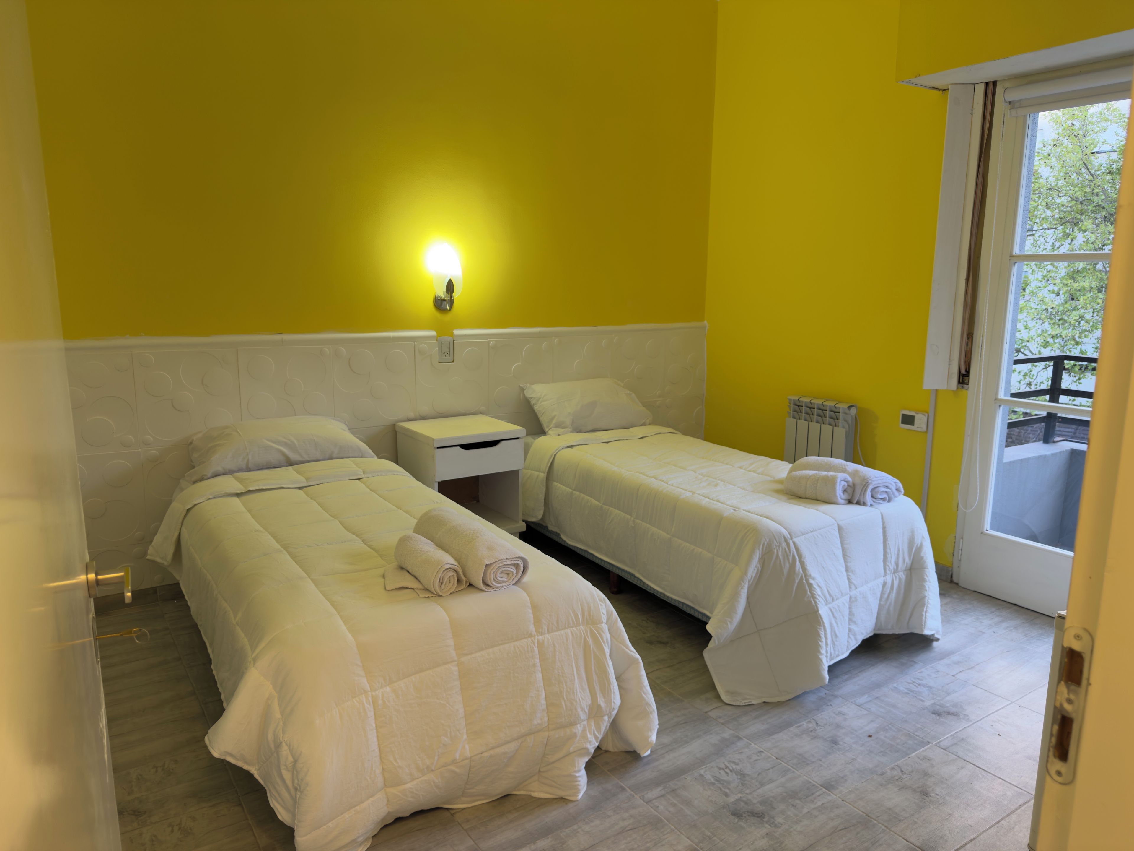 Una stanza con due letti, un comodino, una lampada a parete e una finestra vista.