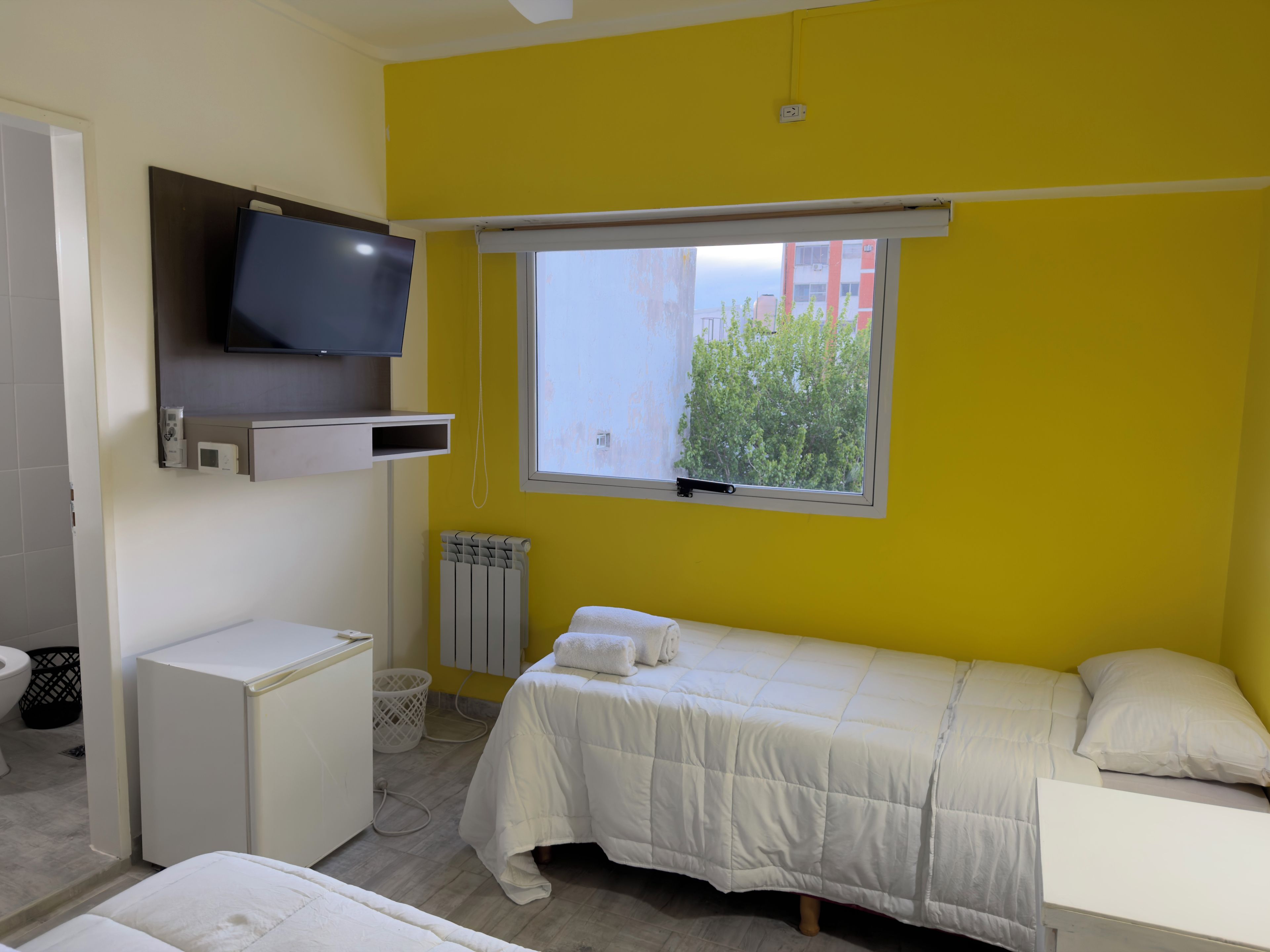 Une chambre à coucher avec un lit, un téléviseur fixé au mur, une fenêtre donnant sur un espace verdoyant et un radiateur.