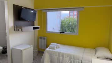 Une chambre à coucher avec un lit, un téléviseur fixé au mur, une fenêtre donnant sur un espace verdoyant et un radiateur.