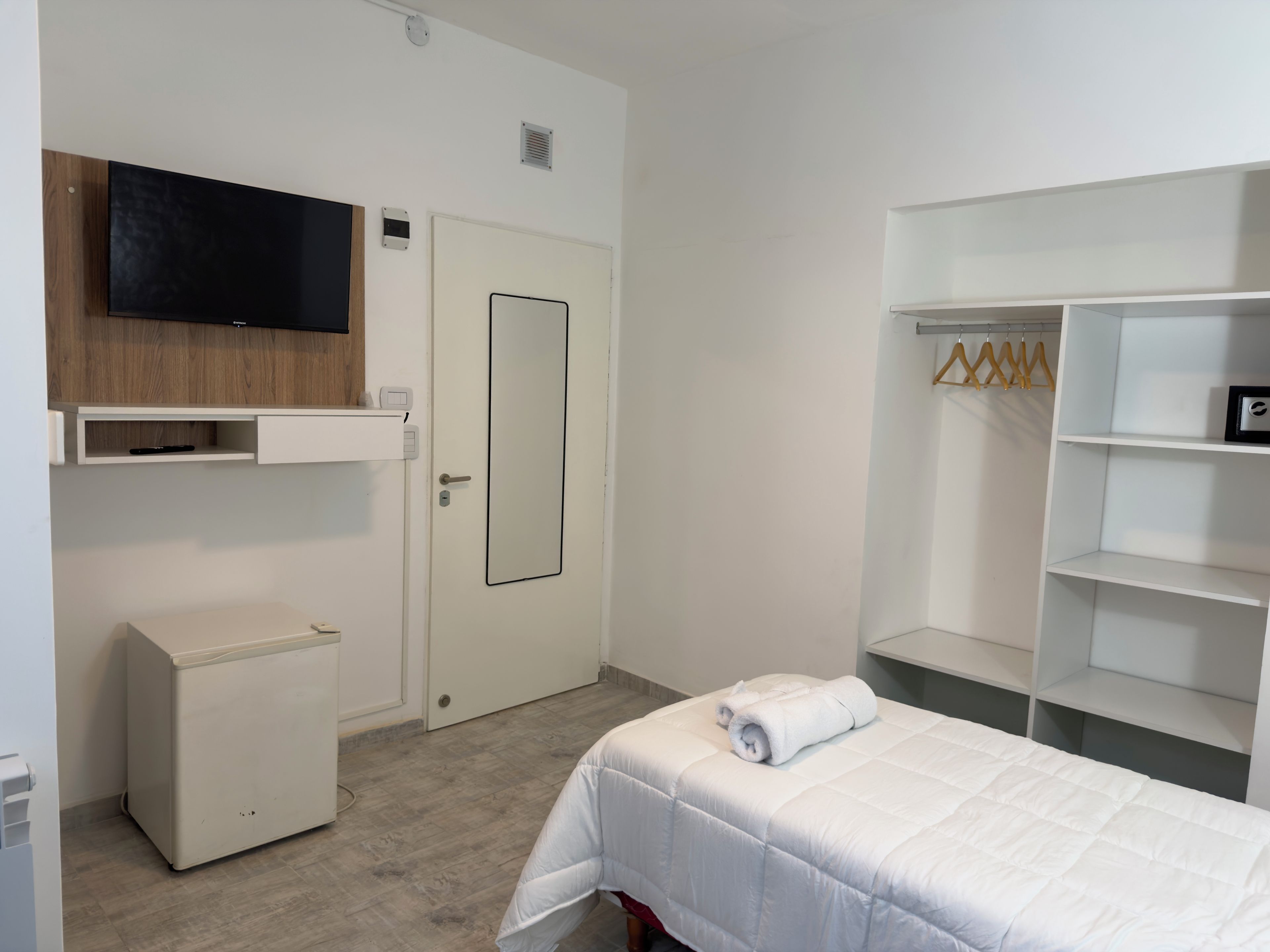 Habitación de hotel con cama, televisor montado en un panel de madera, armario con espacio para colgar y una mesita de noche pequeña.