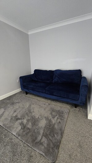 A room with a dark blue sofa against a white wall, a grey carpet, and a white baseboard. - Spacious 3-Bedroom House in Rossendale (Rossendale)