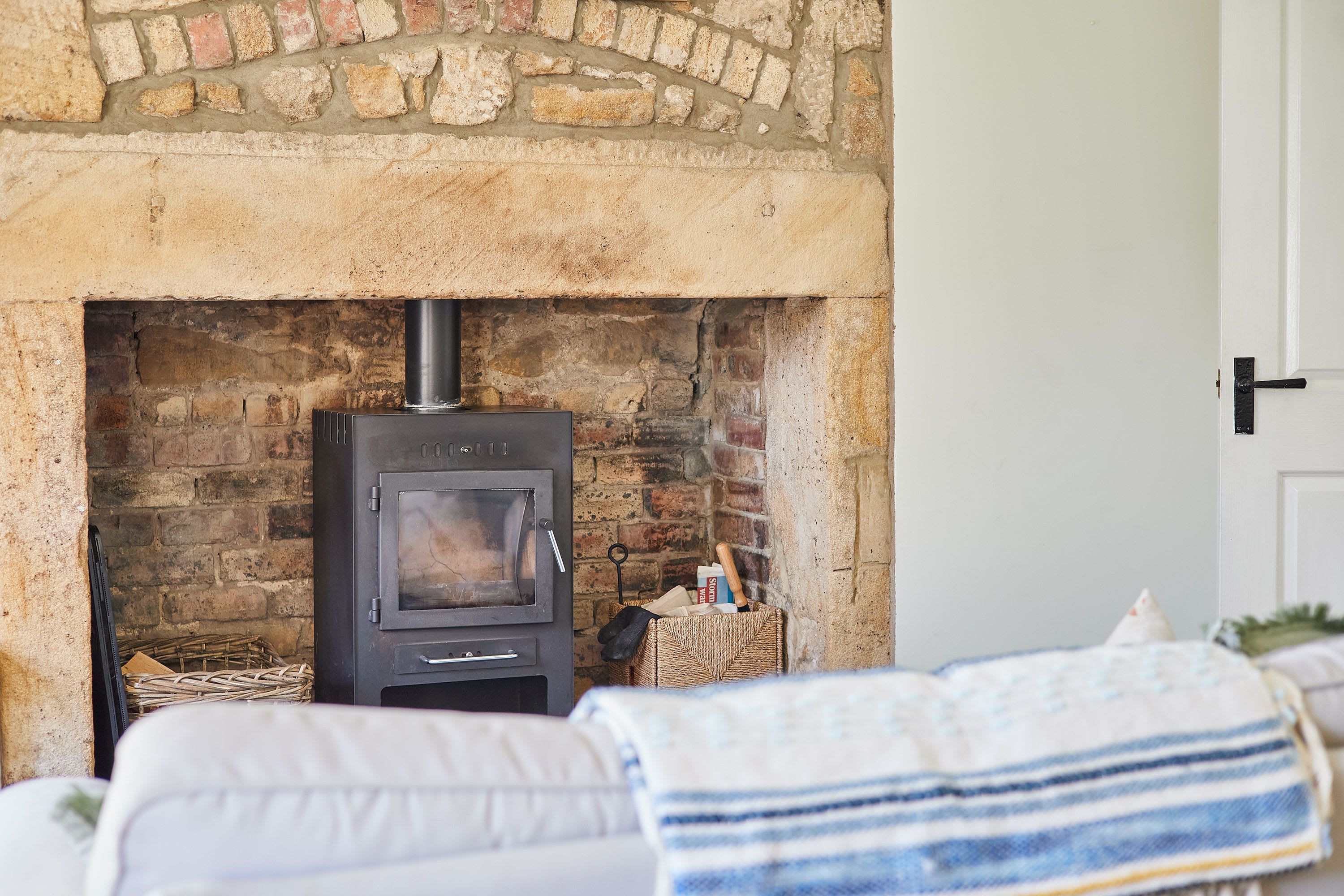 A wood-burning stove in a stone fireplace, a wicker basket with books, and a bed with a striped blanket.